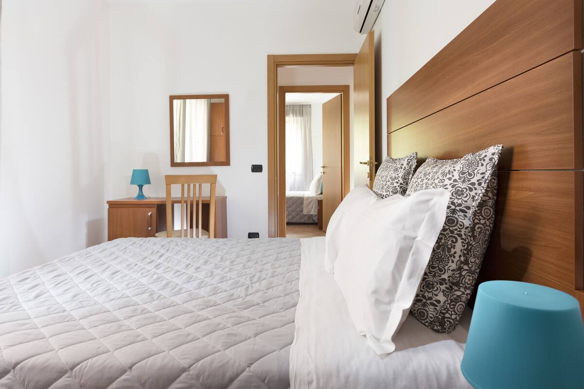 A bedroom with a bed, a wooden headboard, a bedside table with a lamp, and a mirror on the wall.