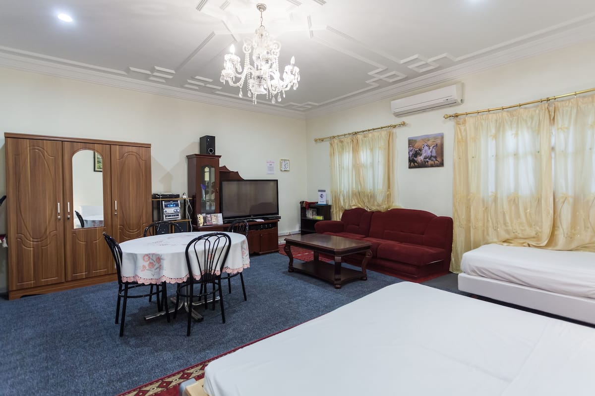 A hotel room with a bed, a red sofa, a dining table with chairs, a TV, a wooden wardrobe, and a chandelier.