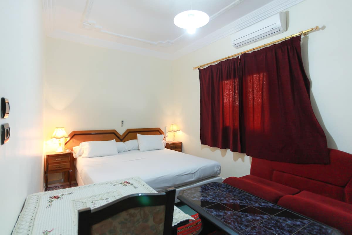 A hotel room with a bed, bedside lamps, a red sofa, a small table, and a wall-mounted air conditioner.