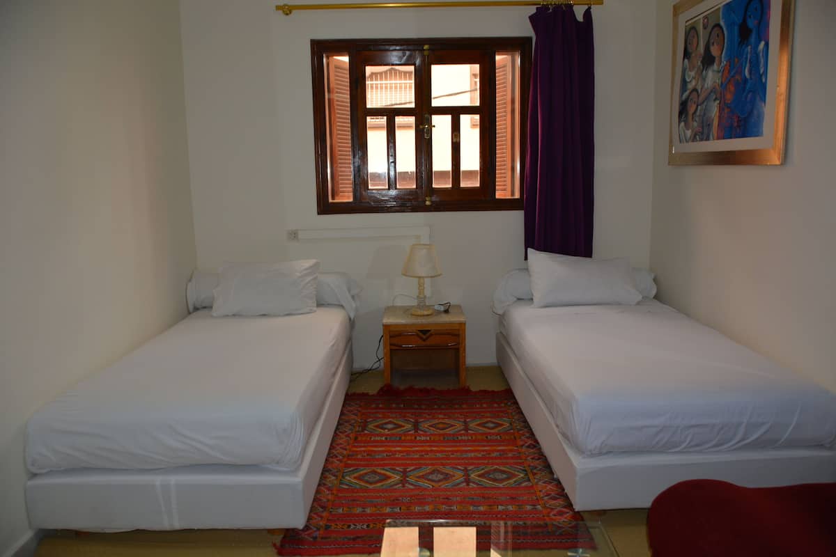A small, clean room with two single beds, a window with shutters, a nightstand with a lamp, and a colorful rug.