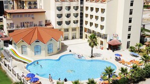 A hotel with a swimming pool surrounded by sun loungers and umbrellas, a children's play area, and a view of mountains in the background. - Larissa Inn (Kemer)