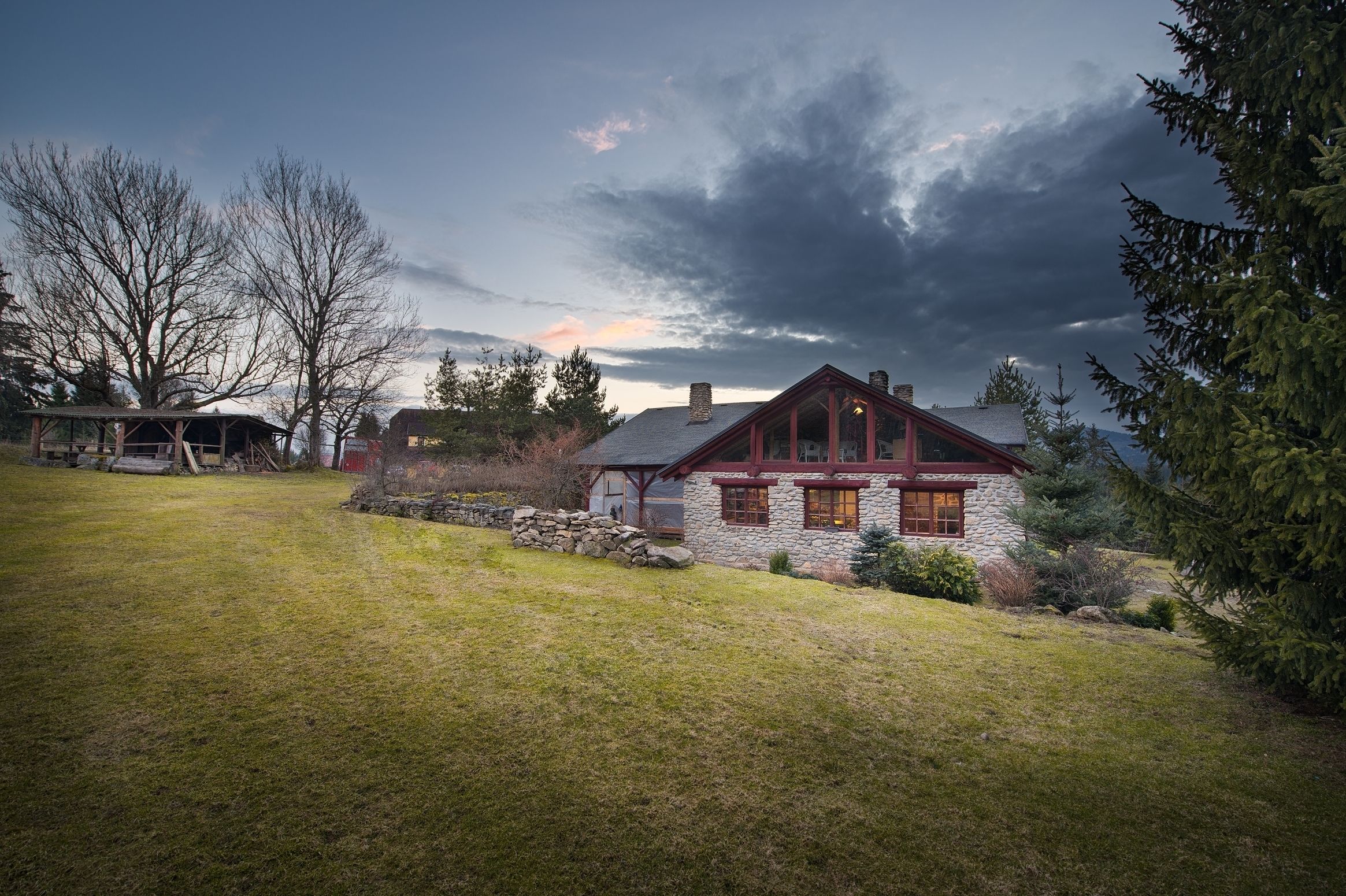 Una casa in pietra con tetto rosso e portico in legno, immersa in un giardino erboso e alberato.