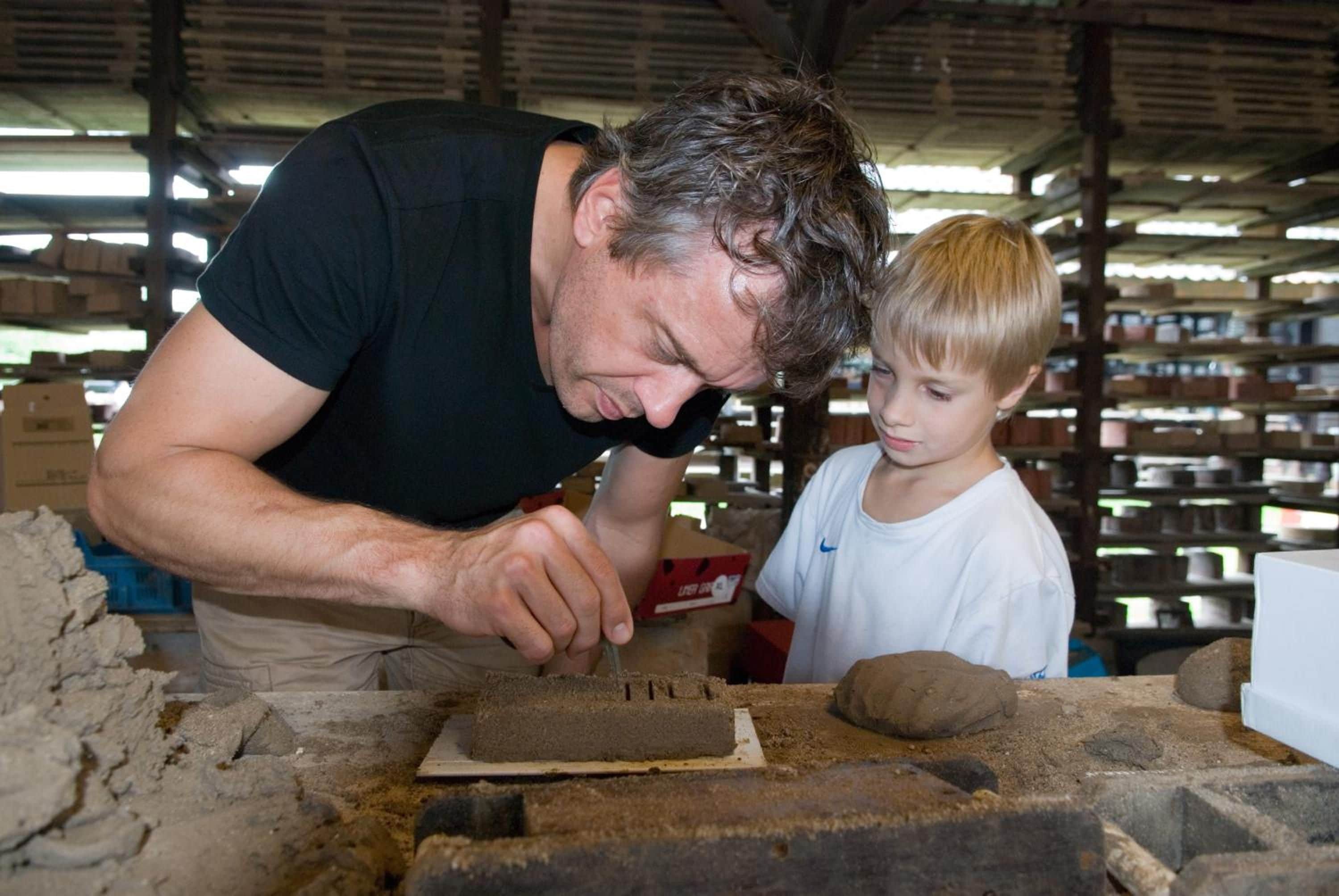 Een man en een kind in een werkplaats, met planken vol dozen op de achtergrond.