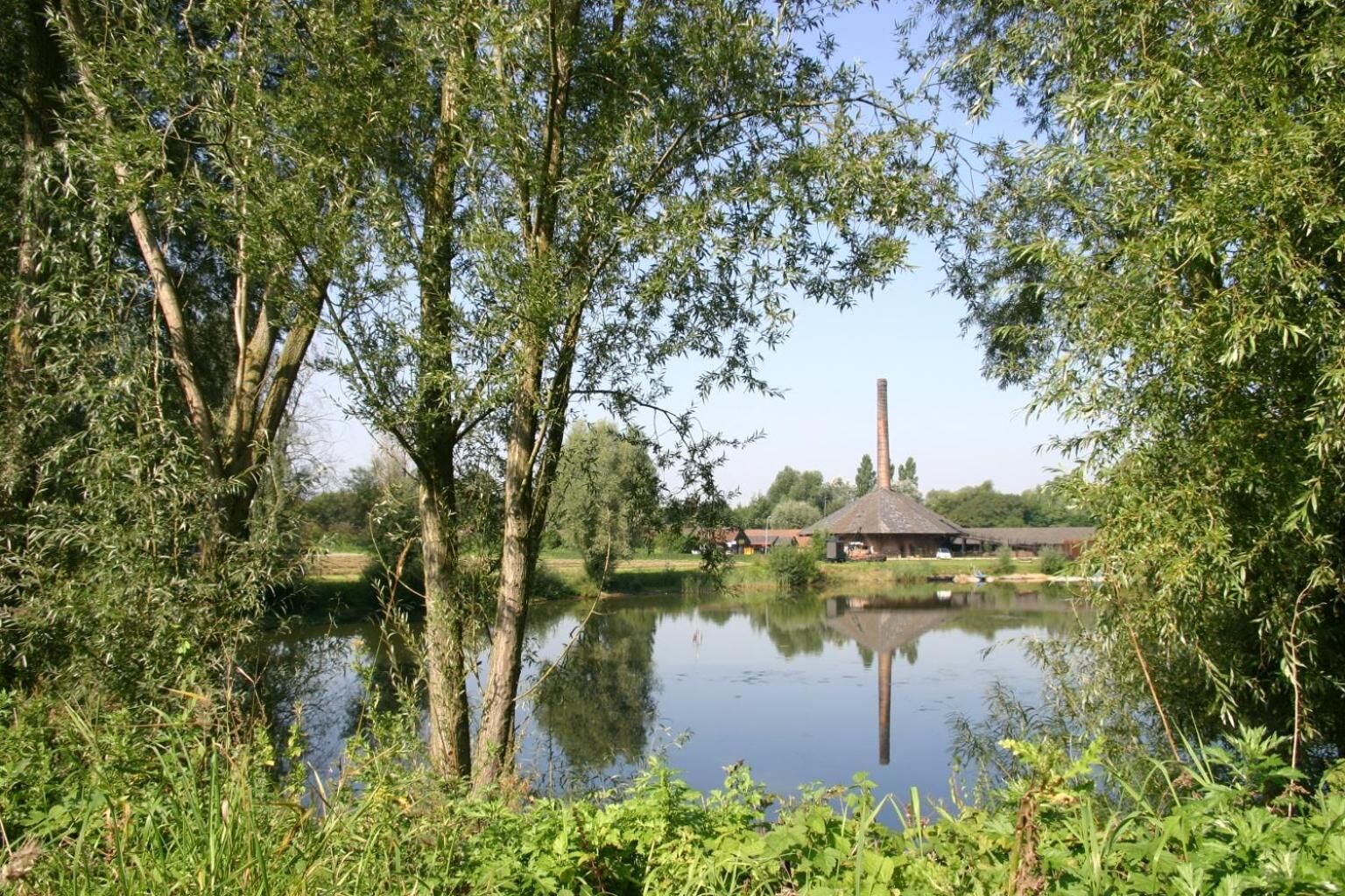 A serene lakeside setting with a traditional thatched-roof building, surrounded by lush greenery and trees reflecting on the calm water.