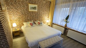 A neatly arranged bedroom with a brick wall, a bed with white bedding, a bench, a bedside table with a lamp, and a window with curtains. - Buitengoed de Panoven (Zevenaar)