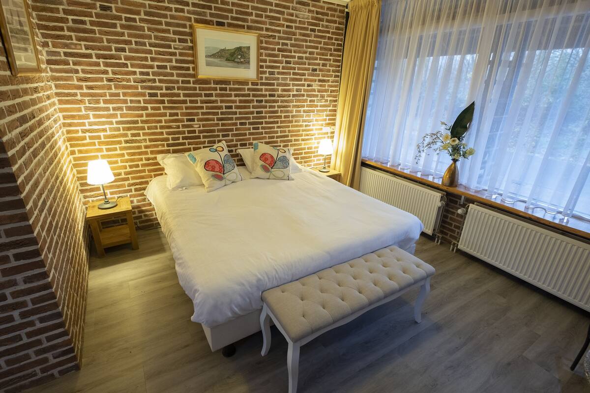Quarto bem arrumado com parede de tijolos, cama com roupa de cama branca, um banco, mesa de cabeceira com abajur e janela com cortinas.