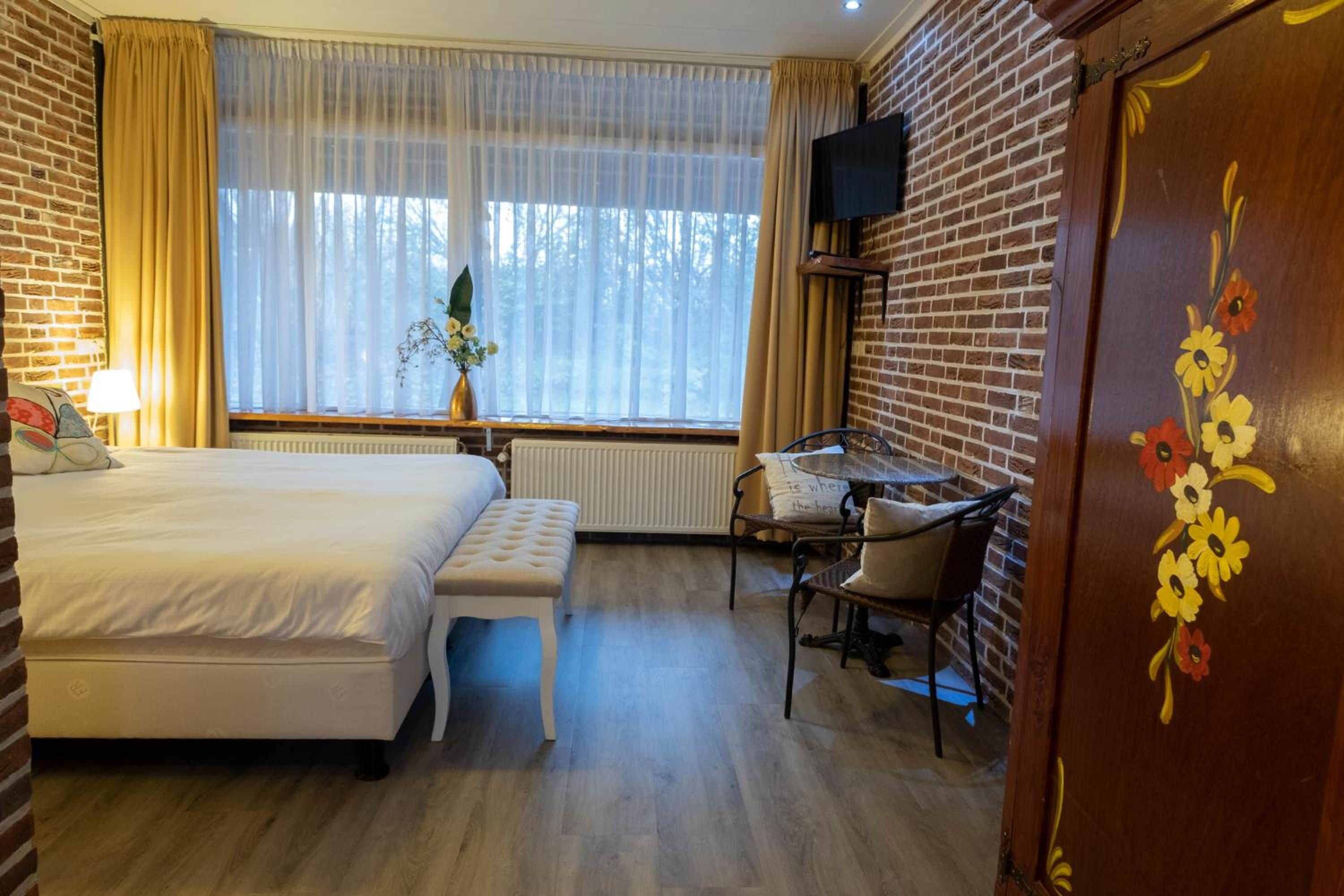 Habitación de hotel con una cama, escritorio, silla, ventana con cortinas y televisor fijado en la pared de ladrillo.