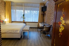 A hotel room with a bed, a desk, a chair, a window with curtains, and a television mounted on the brick wall. - Buitengoed de Panoven (Zevenaar)