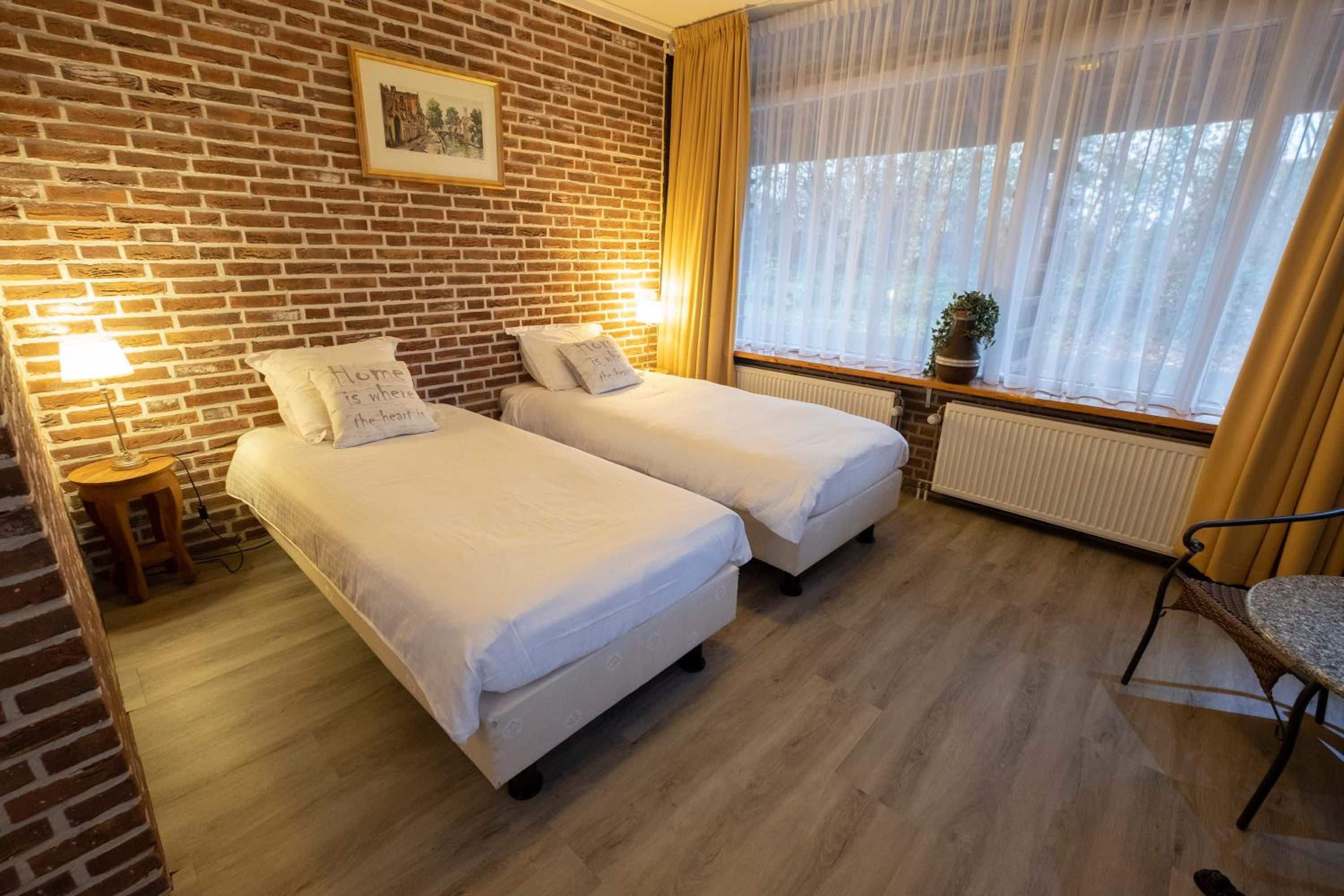 Een kamer met twee eenpersoonsbedden, een houten vloer, een bakstenen muur, een raam met dunne gordijnen en een tafeltje met een lamp.