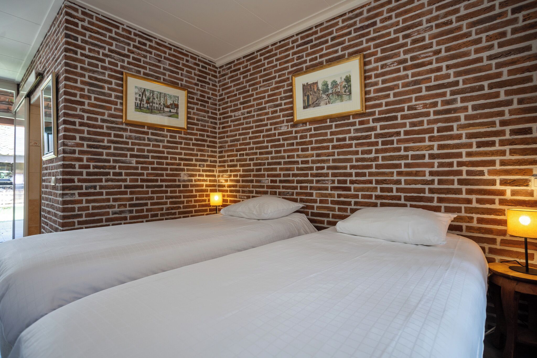 Una habitación con dos camas, paredes de ladrillo, cuadros enmarcados y una mesita de noche con una lámpara.