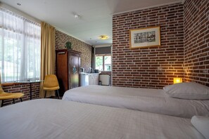 Habitación de hotel con dos camas, un escritorio de madera, una silla, un radiador y una pared de ladrillo con un cuadro enmarcado.
