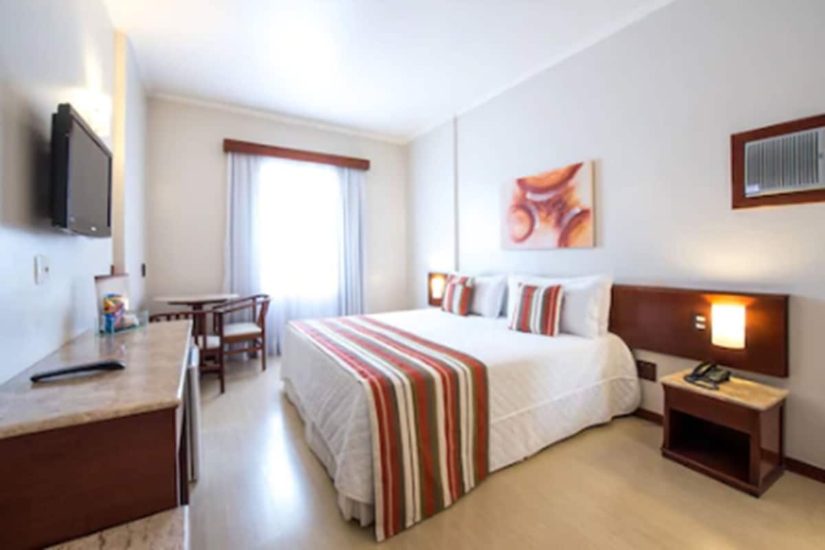 Quarto de hotel com cama, mesa de cabeceira, televisão e secretária com cadeiras.