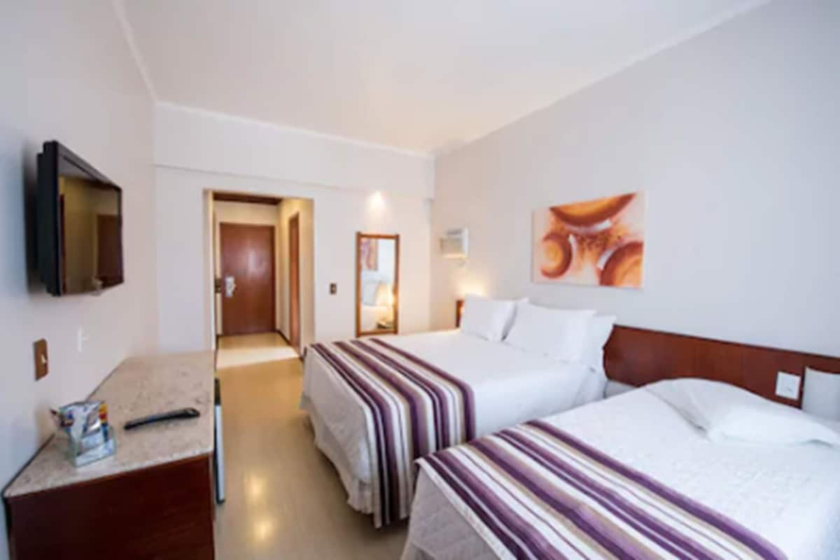 Quarto de hotel com duas camas, uma televisão, um quadro e uma secretária com abajur.