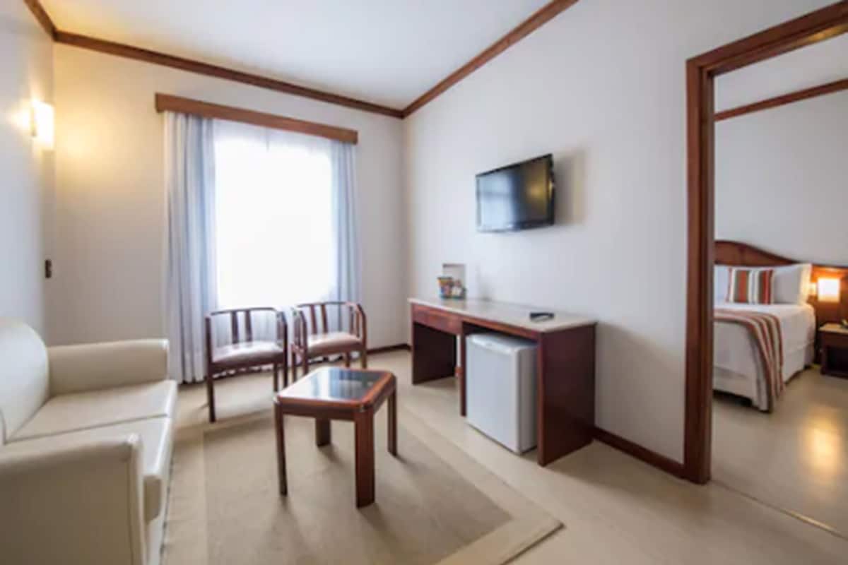 Quarto de hotel com sofá, duas cadeiras, escrivaninha, televisão e cama.