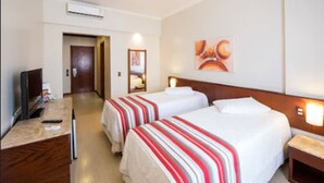 A hotel room with two beds, a desk with a computer, and a TV. There is a door leading to another room and a wall-mounted artwork. - Carlton Plaza Poços de Caldas (Poços de Caldas)