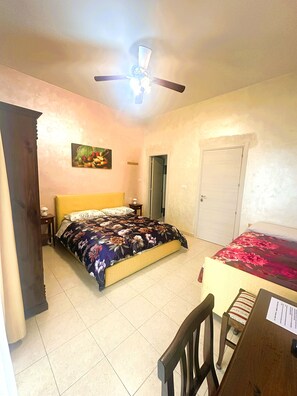 Un dormitorio con cama, escritorio, silla, ventilador de techo, puerta y un cuadro colgado en la pared.