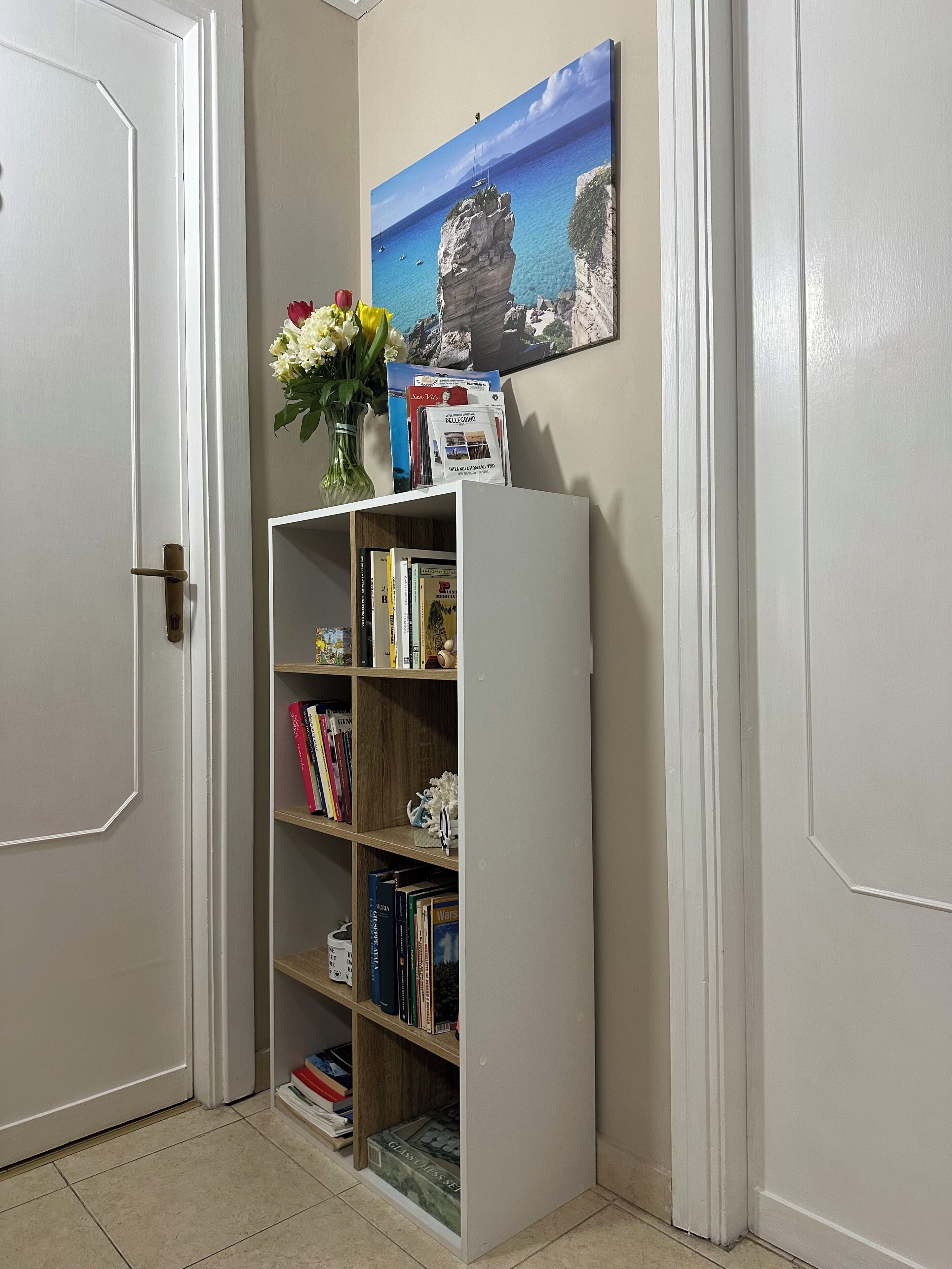 Een boekenplank met boeken en een bloevas, een ingelijst beeld aan de muur, en een deur.