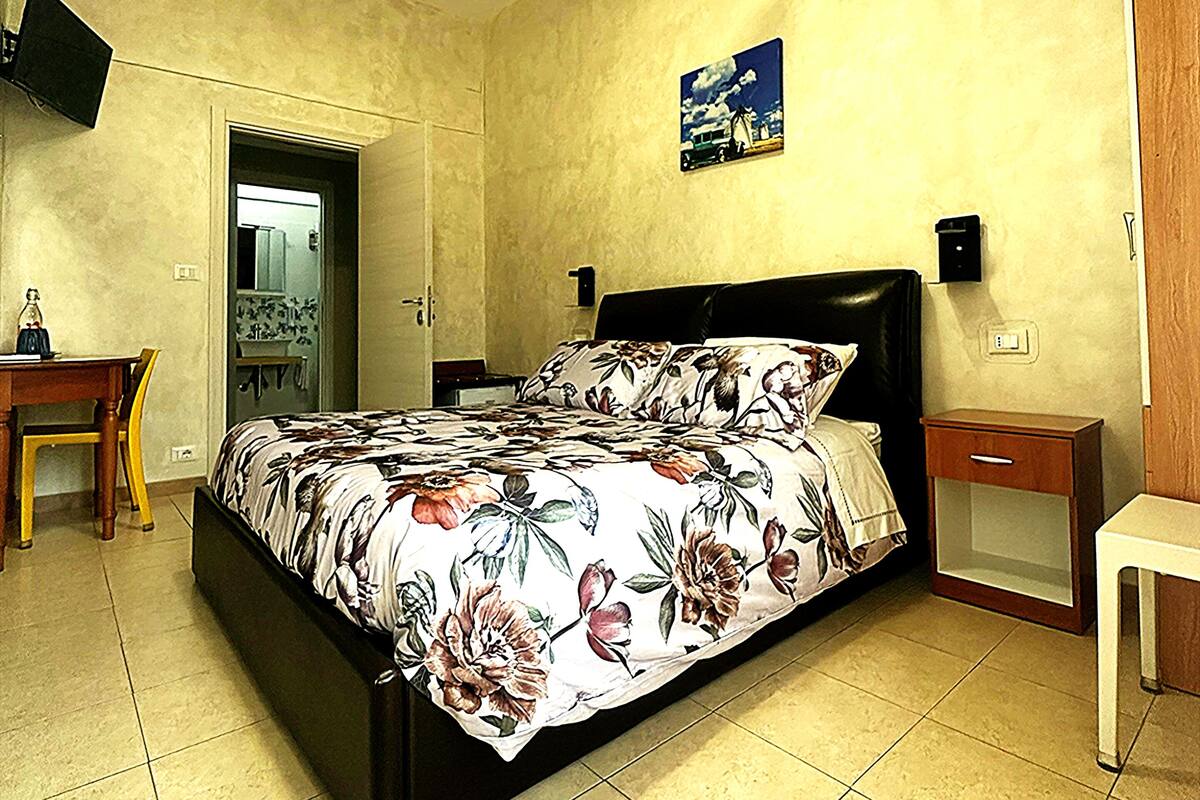 A bedroom with a bed, a ceiling fan, a nightstand, a TV mounted on the wall, and a door leading to another room.