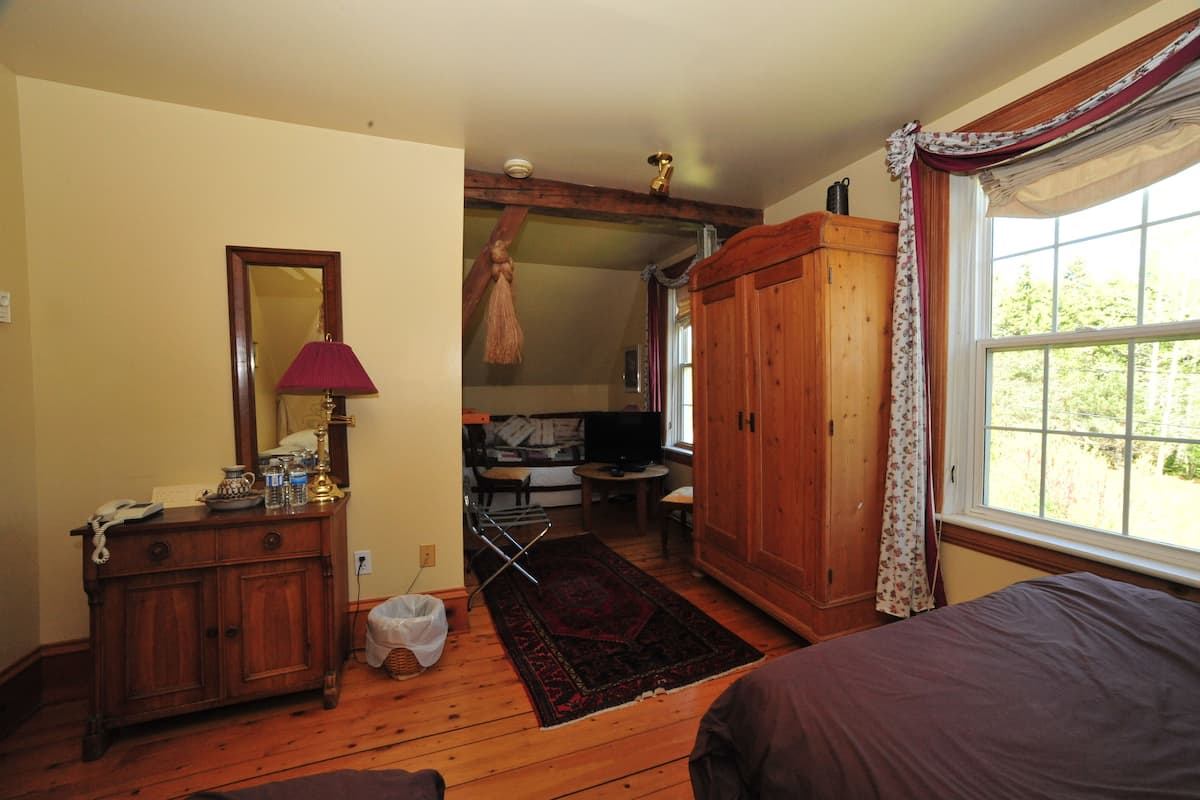 Una habitación con piso de madera, una cama, un aparador con espejo, una lámpara y una ventana con cortinas.