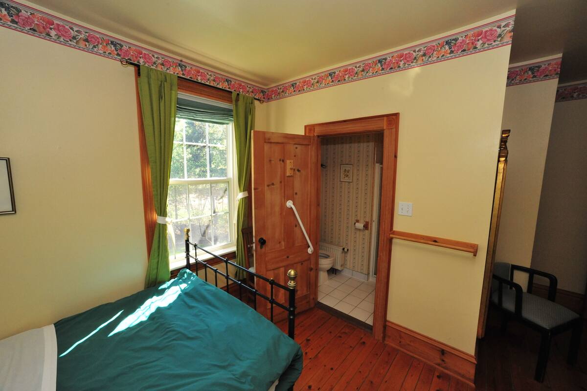 Un dormitorio pequeño y acogedor con una cama individual, una ventana con cortinas, una puerta de madera que da a un baño y una silla.