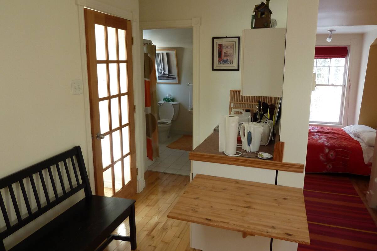 Una habitación con un banco de madera, una mesita pequeña, una cama con colcha roja y una ventana que da a árboles.
