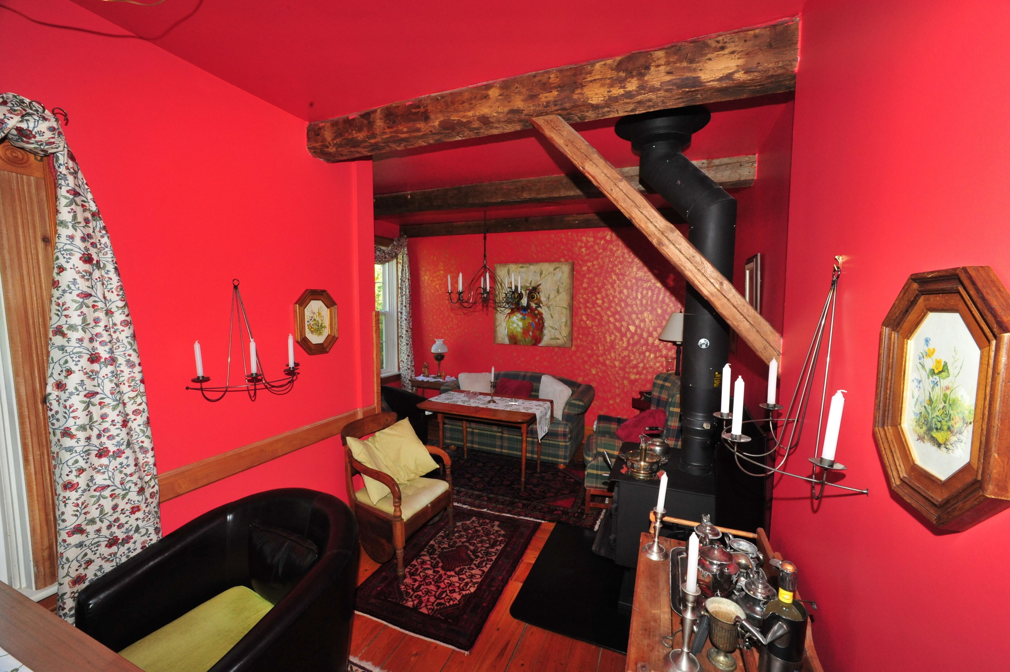 A room with a red wall, a wooden ceiling, a black bathtub, a wooden chair, a table with a floral tablecloth, and a painting on the wall.