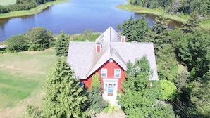 Une maison rouge, dotée d’un toit à pignon, entourée d’une végétation luxuriante et bordée d’une vaste étendue d’eau.
