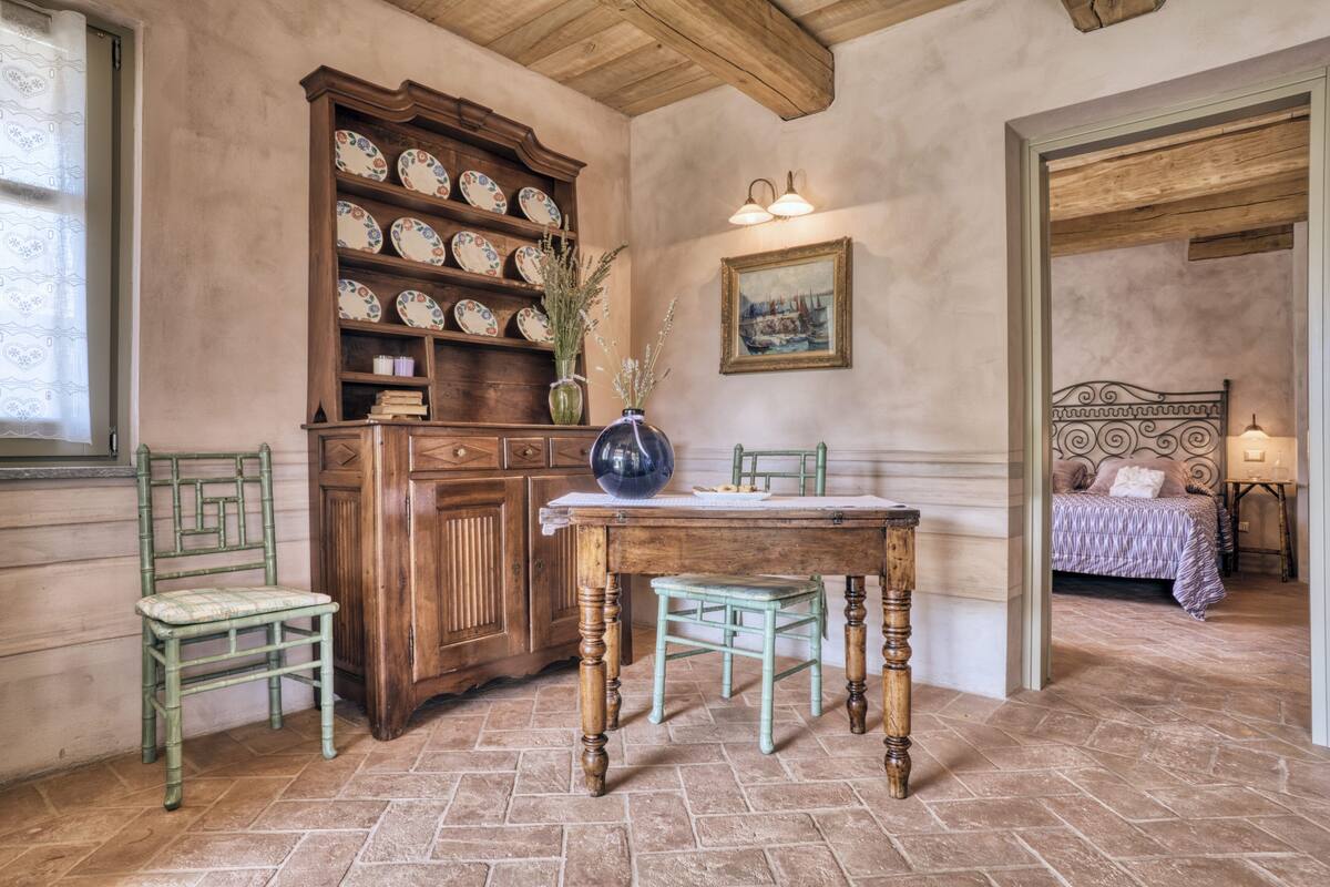 A rustic room with a wooden dining table, a green chair, a cabinet with decorative plates, a vase with flowers, and a bed with a patterned bedspread.