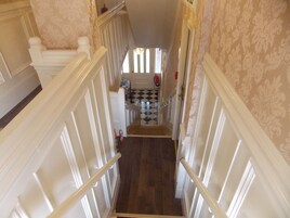 Una escalera estrecha con barandillas blancas y papel tapiz estampado, que lleva a una habitación con suelo a cuadros.