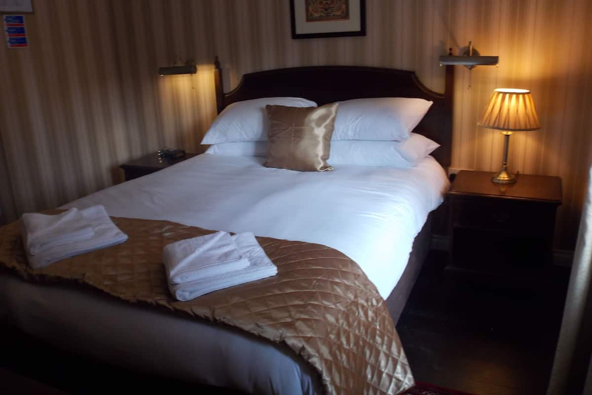 Una cama bien hecha con sábanas blancas y un plaid marrón, una lámpara de noche y un cuadro en la pared.