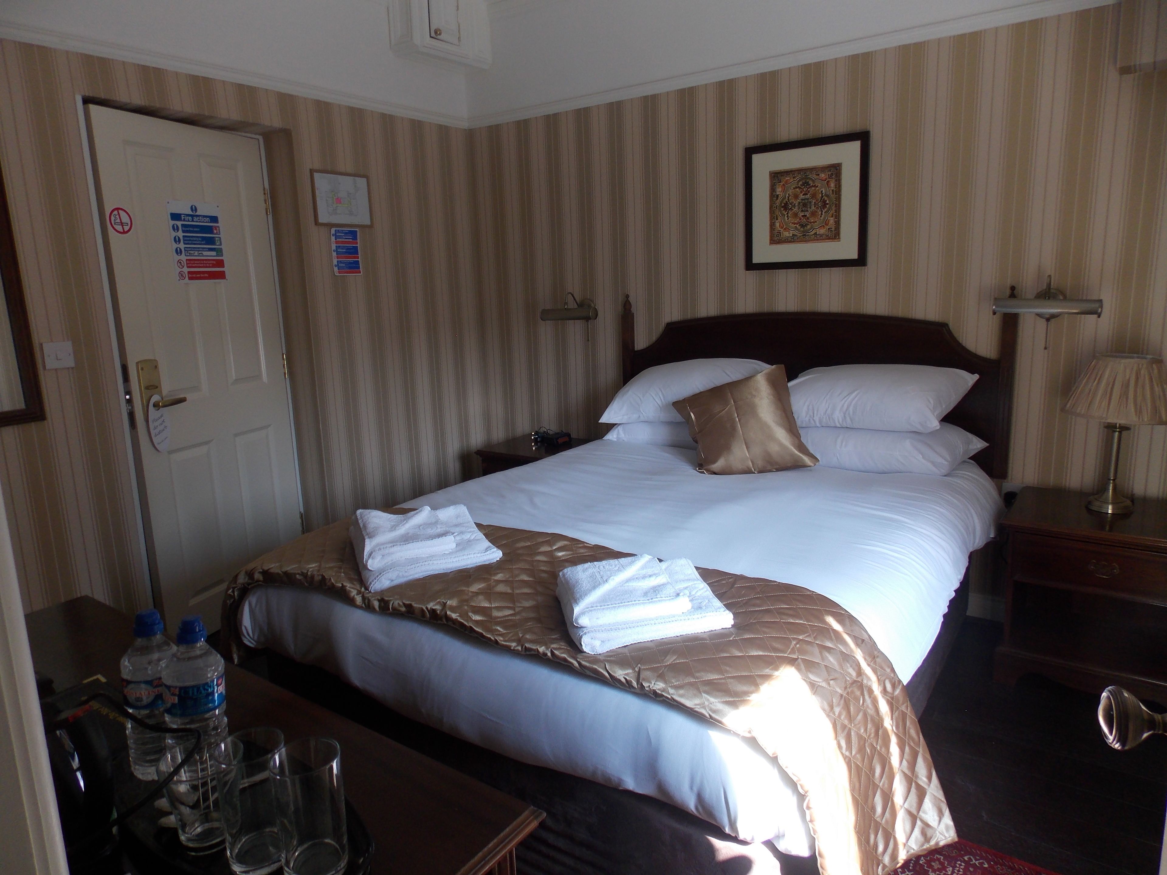 A hotel room with a bed, a nightstand, a lamp, a framed picture on the wall, and a door with a fire action sign.