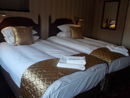 Quarto de hotel com duas camas, cada uma com colcha matelassada dourada e roupa de cama branca. Há duas luminárias de cabeceira e um espelho na parede.