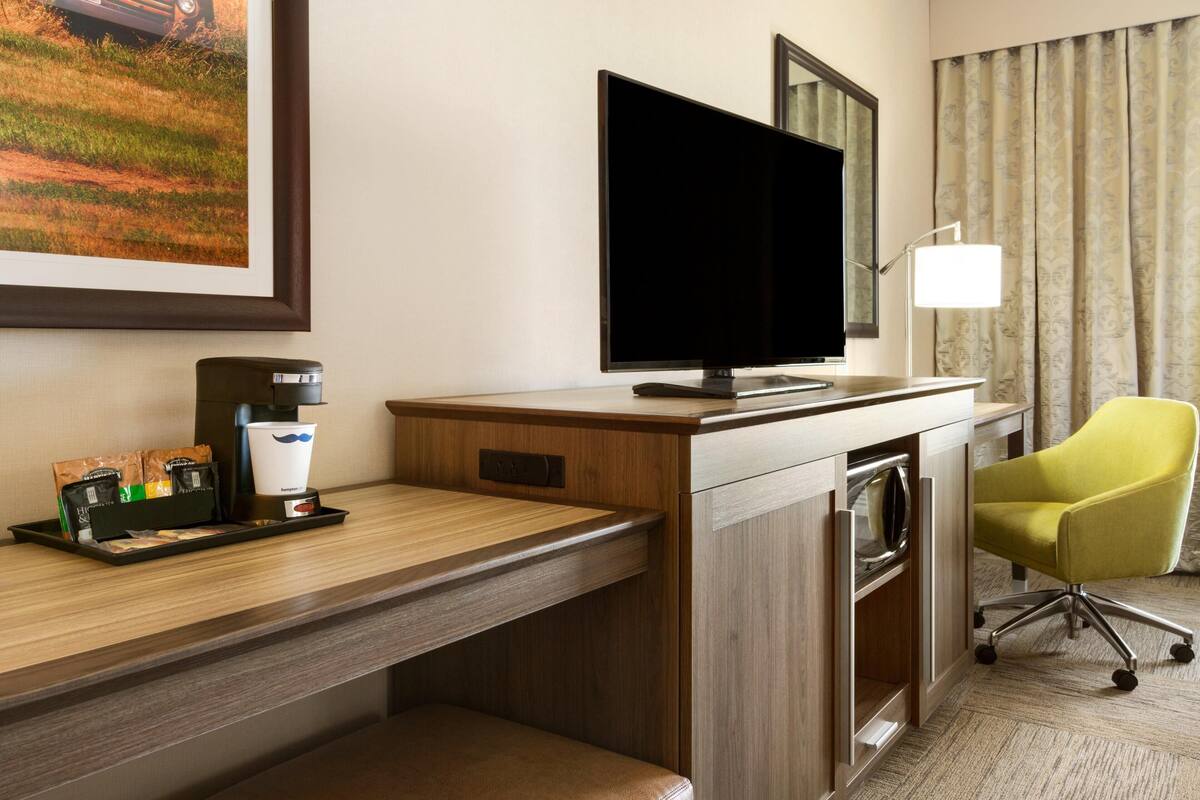 A hotel room with a wooden desk, a flat-screen TV, a coffee maker, a trash bin, a chair, and a large window with curtains.