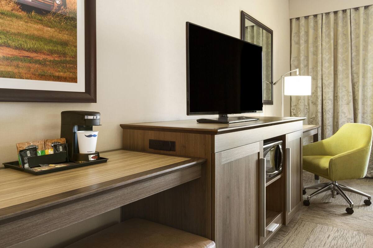 A hotel room with a wooden desk, a flat-screen TV, a coffee maker, a tray with coffee packets, a chair, and a large window with curtains.