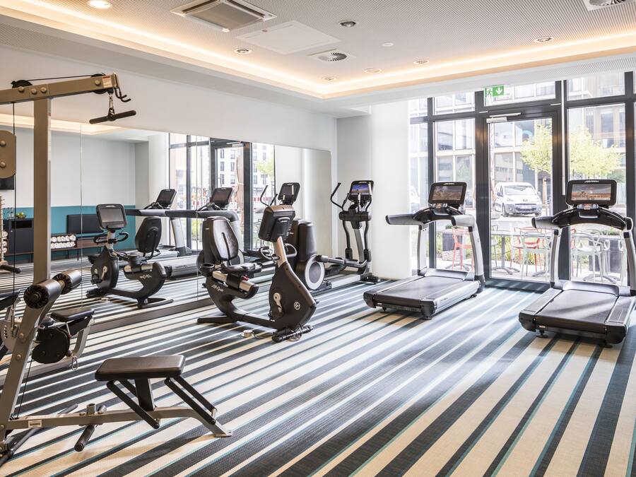 A modern gym with various exercise equipment, large windows, and a striped floor.