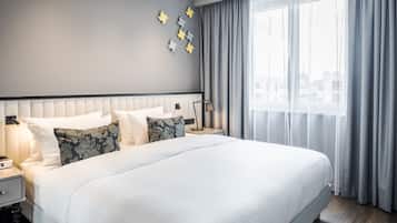 A modern hotel room with a large bed, a nightstand with a lamp, and a view of the city through sheer curtains.