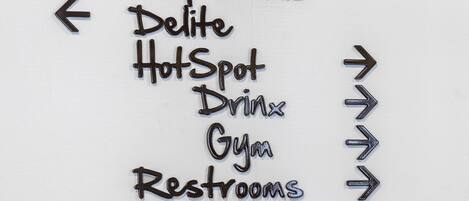 Un letrero direccional con flechas que indican diferentes servicios, como 'Pow-Wow', 'Capricious', 'Delite', 'Hot Spot', 'Bebidas', 'Gimnasio' y 'Baños'.