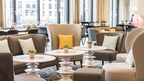A modern hotel lobby with comfortable seating, a dining area, and large windows offering city views.