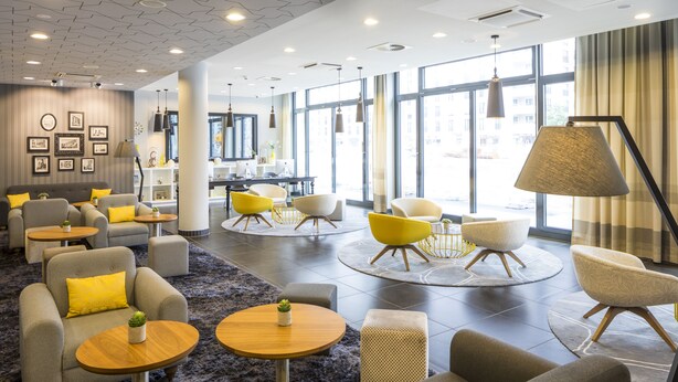 A modern hotel lobby with a mix of seating areas, including sofas and armchairs, arranged around coffee tables and a central dining table. Large windows provide natural light and a view of the cityscape.
