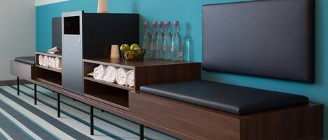 A modern hotel room with a long bench, a wall-mounted TV, a shelf with towels, a vase with an orchid, and a basket of apples.