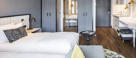 Una habitación de hotel moderna con una cama grande, una zona de estar con sofá, una mesita y vistas al baño y la cocina.
