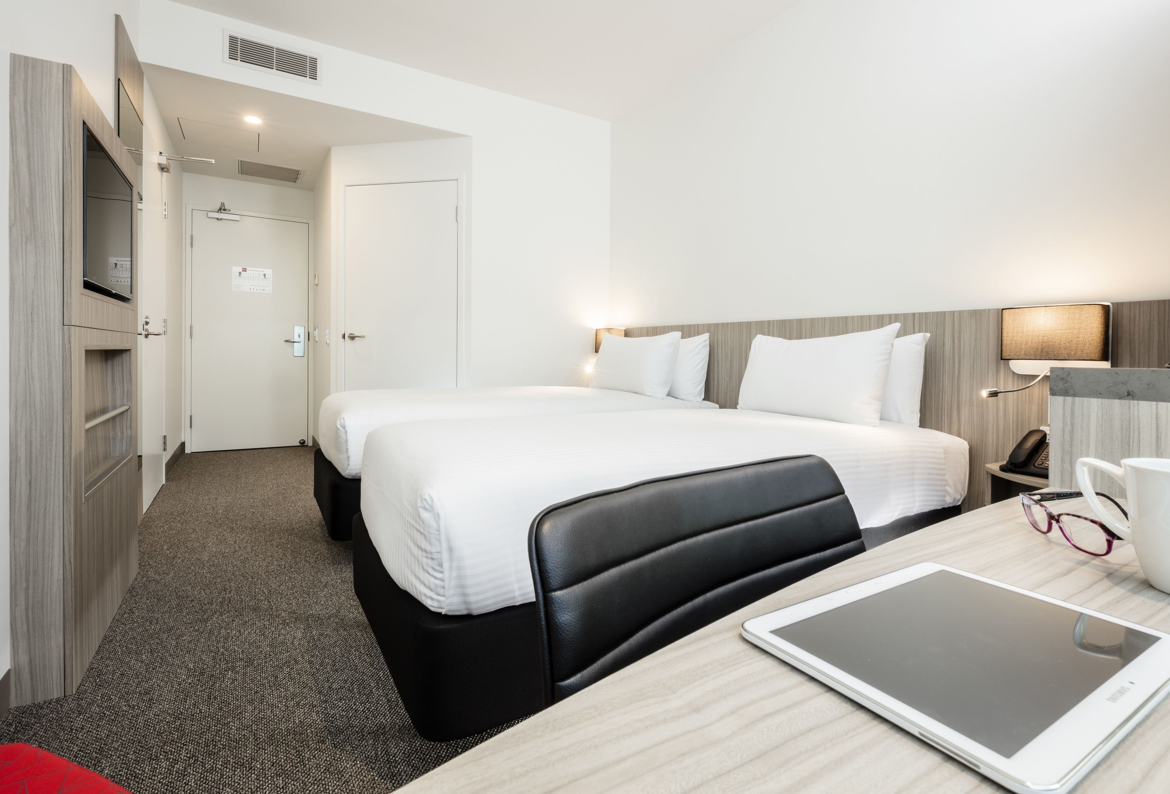 A hotel room with two beds, a desk with a tablet, a chair, a bedside table with a lamp, and a door leading to a bathroom.