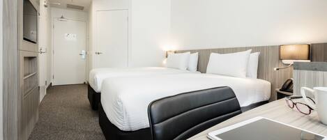 A hotel room with two beds, a desk with a tablet, a chair, a bedside table with a lamp, and a door leading to a bathroom.