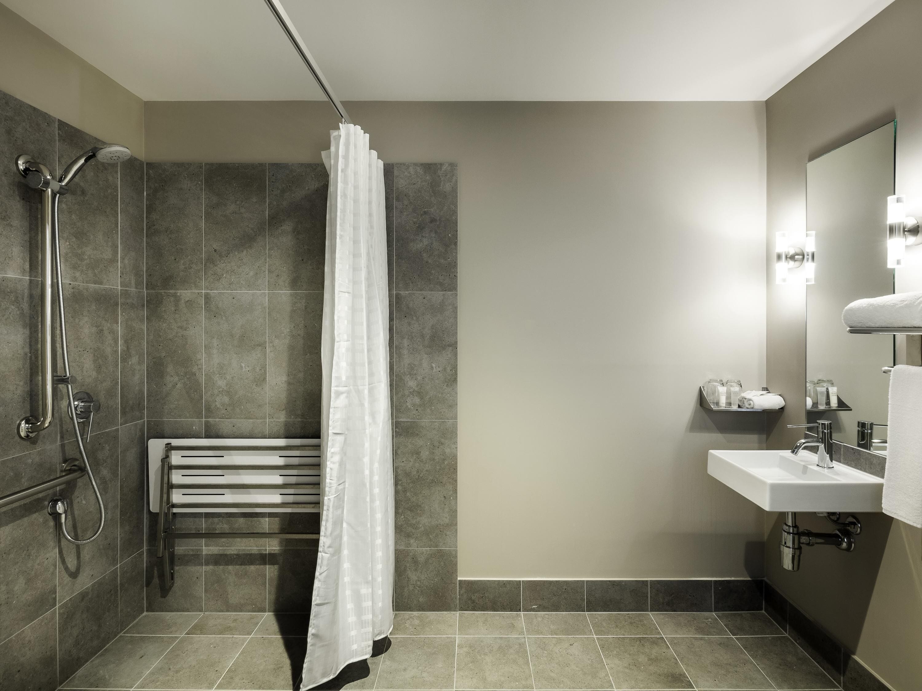 A modern bathroom with a shower, a bench, a sink, and a mirror.