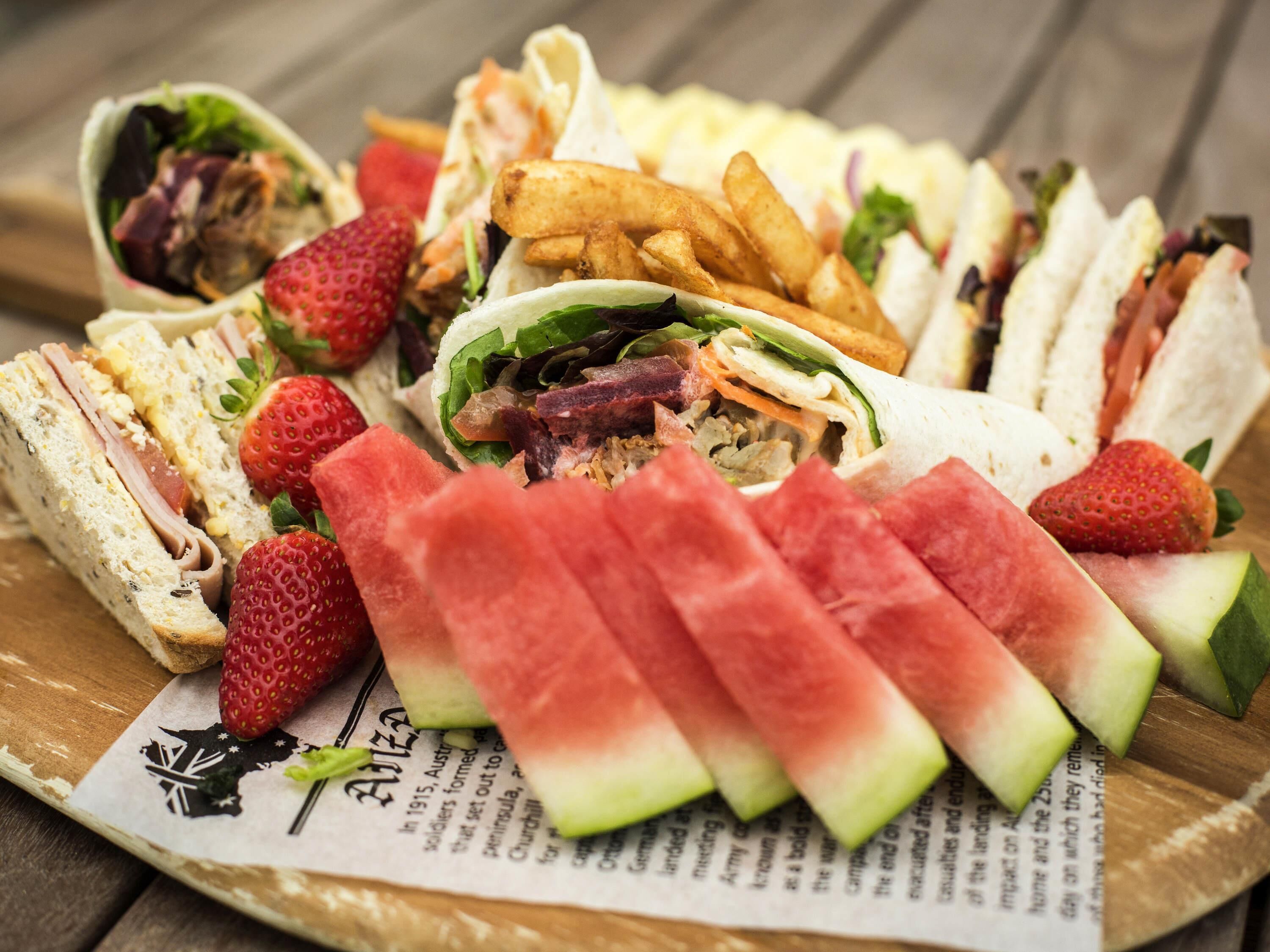 A variety of sandwiches, including wraps and burgers, alongside sliced watermelon and strawberries on a wooden surface.