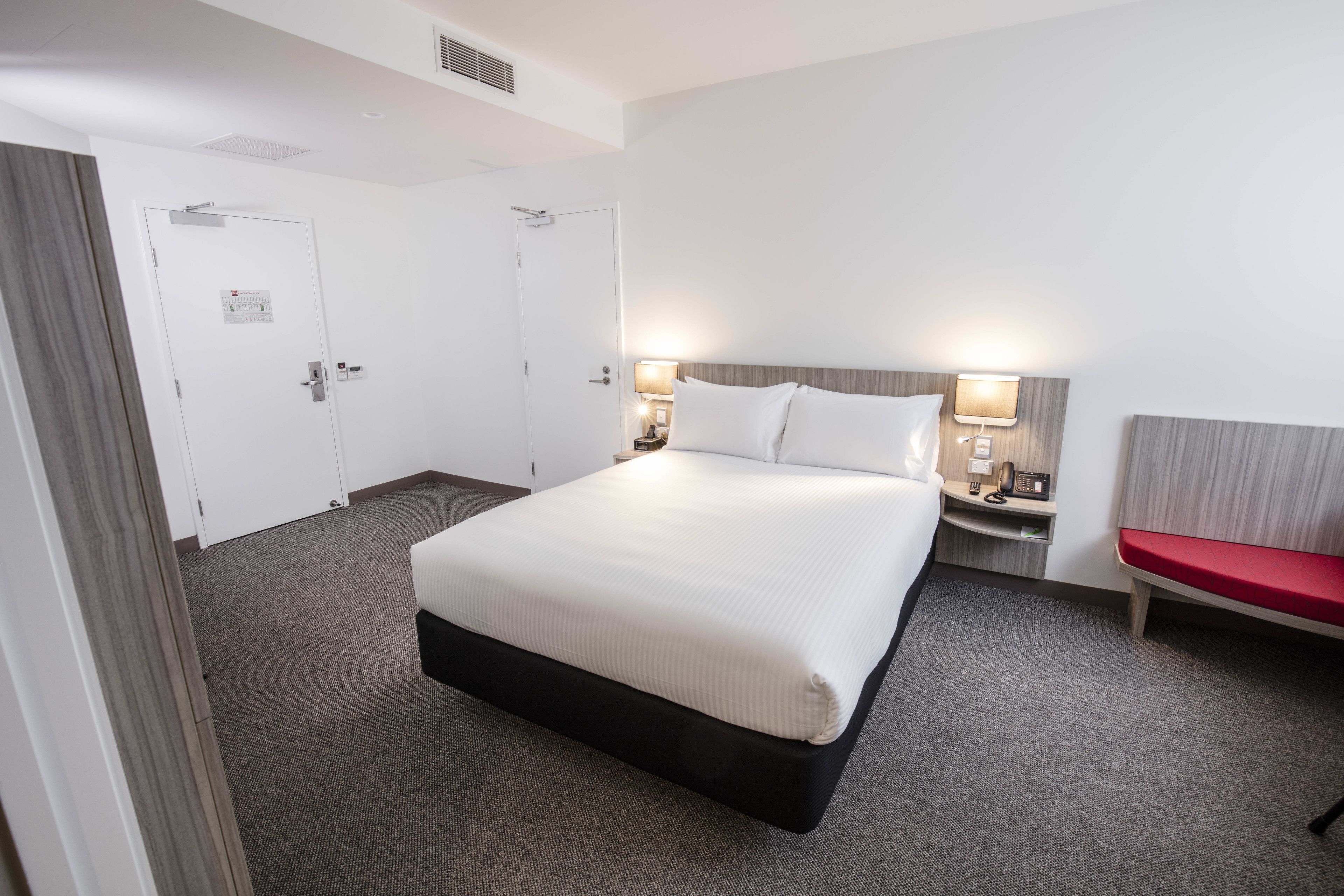 A hotel room with a large bed, two bedside tables with lamps, a red bench, and two doors.