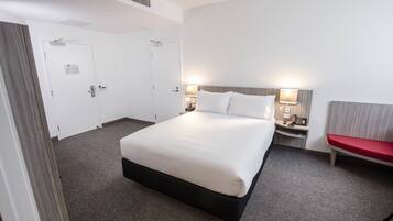 A hotel room with a large bed, two bedside tables with lamps, a red bench, and two doors.