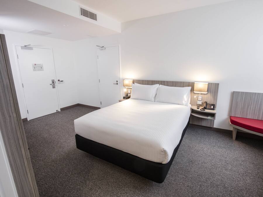 A hotel room with a large bed, two bedside tables with lamps, a red bench, and two doors.