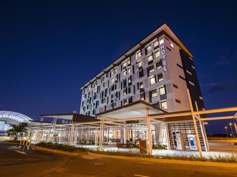 Un hotel moderno con fachada de vidrio, entrada cubierta y estacionamiento visible.