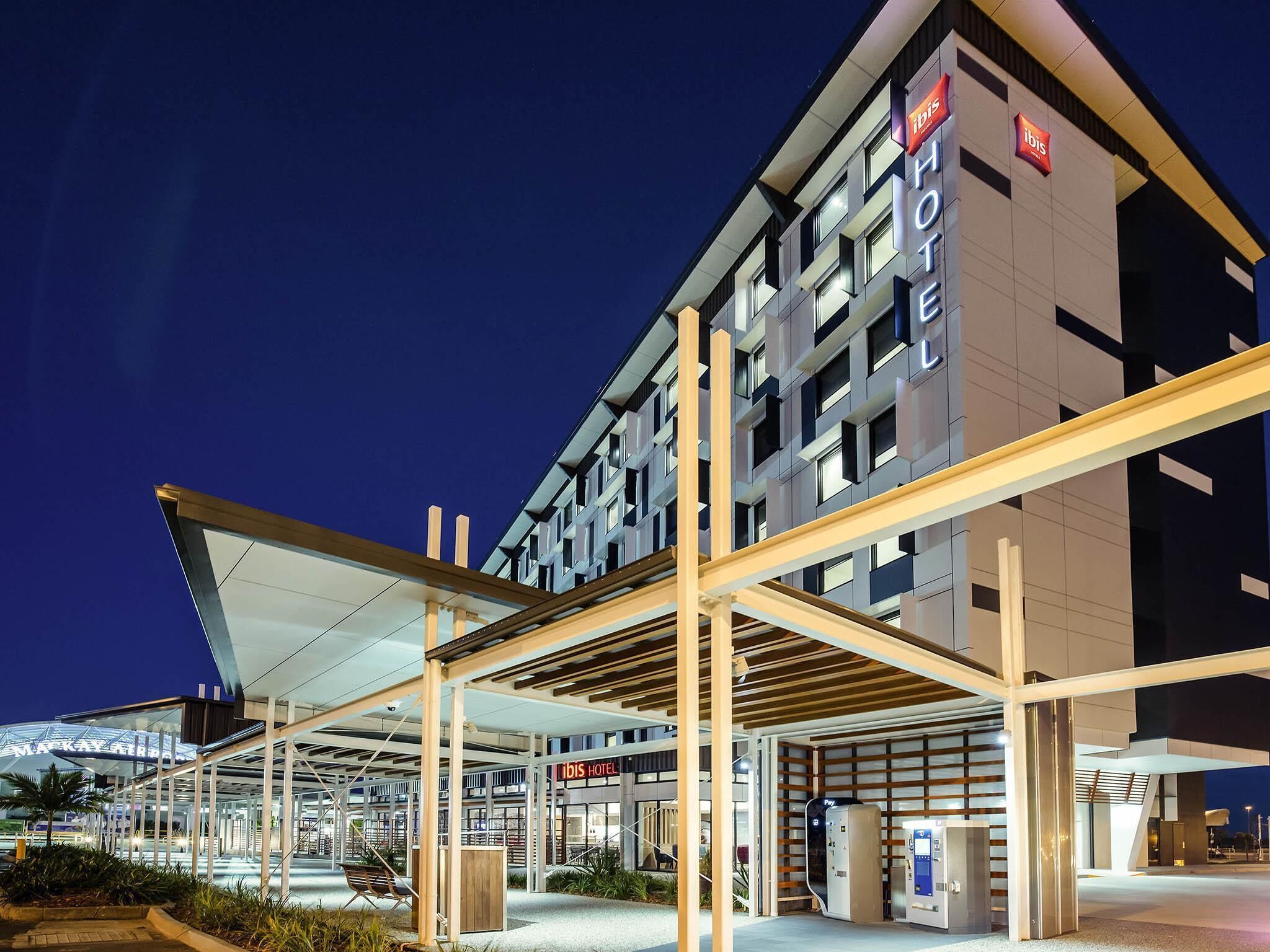 A modern hotel with a prominent 'ibis' sign, a covered entrance area, and a visible payment machine.